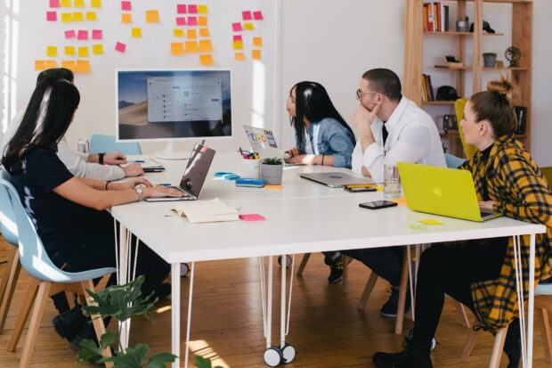 photo of people around table with laptops all conversing whilst looking at screen. Lots of post its on wall