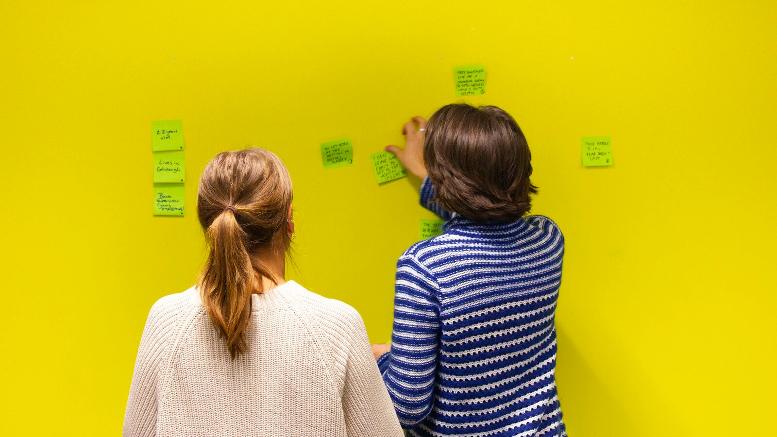 Photograph of two people sticking green post its with writing onto a bright yellow wall. The people appear to be in discussion. 