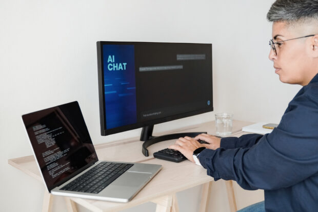 A person seated at a desk with two laptops open in front of them. The person is interacting with an AI chatbot on one of the laptops, while the other laptop displays an open document.