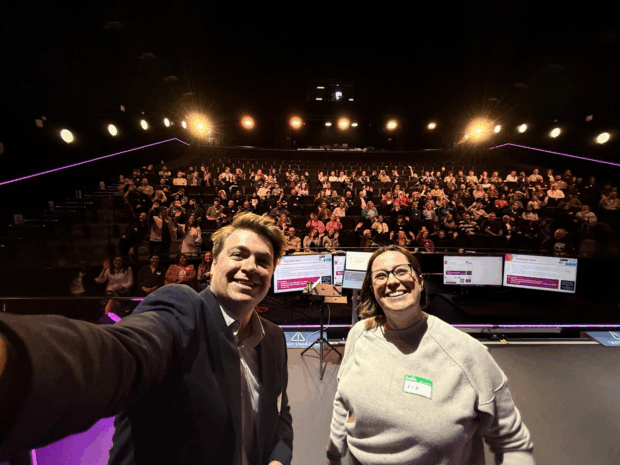 Two presenters stand at the front of a large auditorium taking a selfie with the audience behind them. The man on the left is smiling and holding the camera out, while the woman on the right, wearing glasses and a grey jumper with a “Liz” name sticker, is also smiling. Behind them, rows of attendees sit in tiered seating, waving and looking toward the stage. Large screens with presentation slides are visible in front of the audience, and stage lights illuminate the scene.